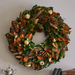 Decorative wreath with green leaves, pinecones, and gold balls on a plain wall.