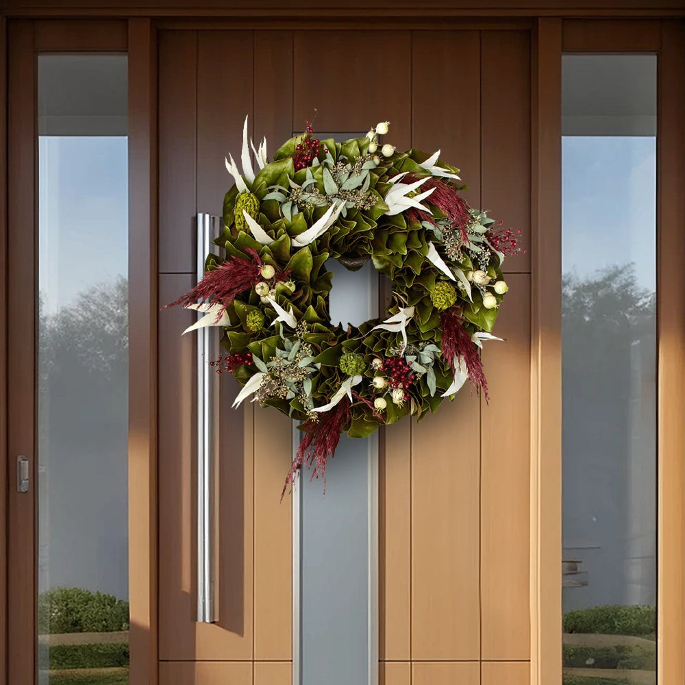 Decorative wreath of green magnolia leaves, white leaves, red accents, and dried flowers on a modern wooden front door with glass panels.
