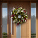 Decorative wreath of green magnolia leaves, white leaves, red accents, and dried flowers on a modern wooden front door with glass panels.