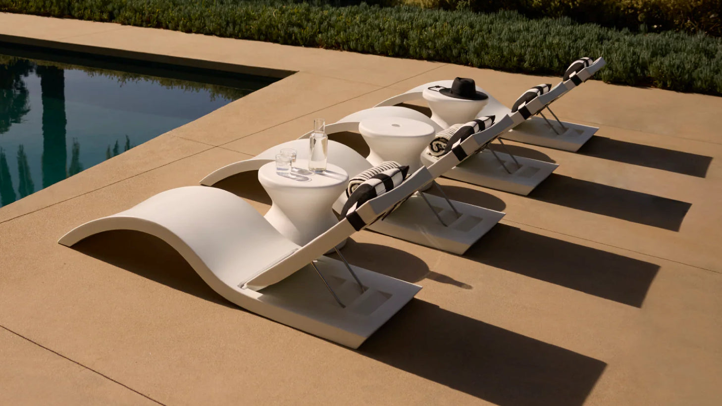 Modern white pool chaise lounges with striped cushions and pedestal side tables beside a luxury outdoor pool.