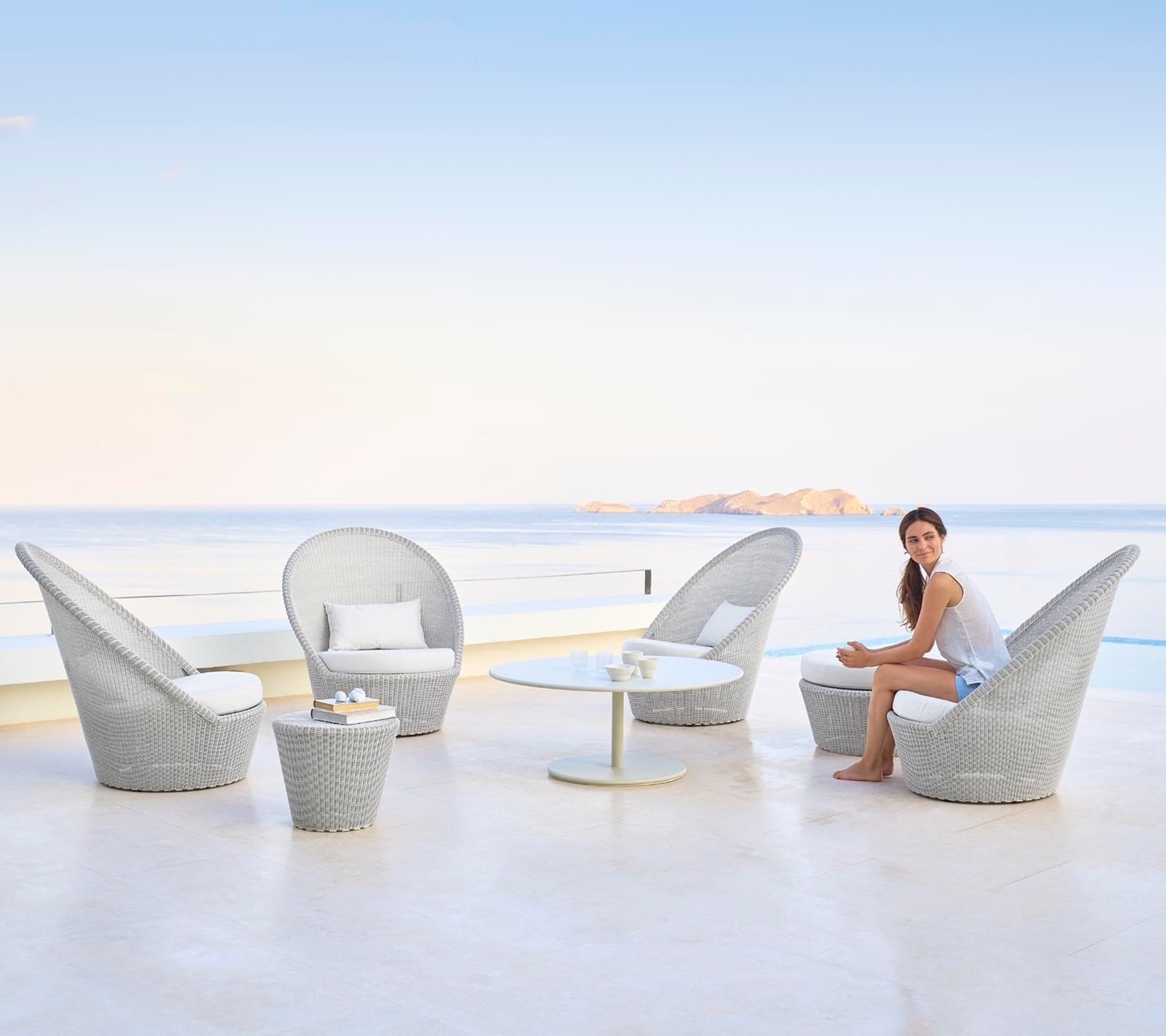 Boxhill's Kingston Sunchair Lounge with Wheels lifestyle image with a woman sitting down on it and Kingston Outdoor Footstool | Side Table and white round table at seafront