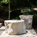 Log Roman Outdoor Stone Side Table holding a potted plant beside a matching low table in a leafy garden.