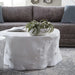 A round, textured white coffee table styled in a living room setting with a gray sofa, blue pillows, and a potted air plant on top.
