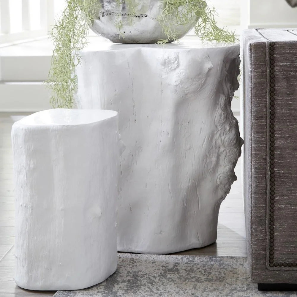 Two white faux log side tables in a modern living room setting, with one smaller cylindrical table in front of a larger rectangular table topped with a hanging plant.