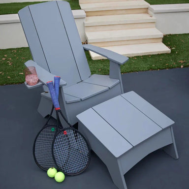 A light gray outdoor Adirondack chair with a matching ottoman on a patio, with two tennis rackets and two tennis balls placed in front. Grass and steps are visible in the background.