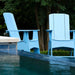 Two light blue outdoor Adirondack chairs placed beside a swimming pool, with trees and shrubs in the background.