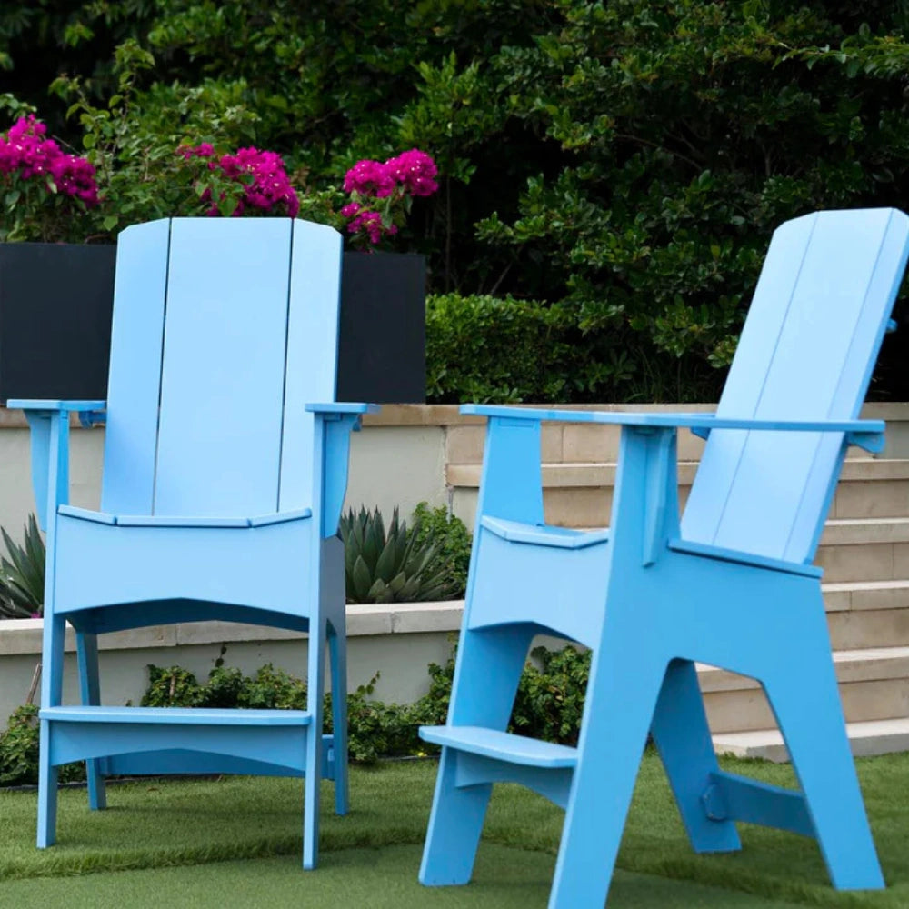 Two bright blue outdoor Adirondack chairs placed on green grass in front of a low wall, with potted pink flowers and dense green foliage in the background.