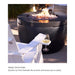 A white outdoor Adirondack chair with a swivel-out drink holder attached to the armrest, holding a wine glass. In the background is a black round fire pit with flames burning inside, set in an outdoor patio environment.