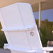 A close-up of a white outdoor patio chair showing the backrest and armrests, with the brand name 'FLEEDGE' embossed on the upper panel. The background is a blurred outdoor patio setting with a canopy and greenery.