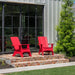 Two bright red outdoor Adirondack chairs placed on a brick patio in front of a house with large black-framed glass doors and green landscaping.