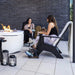 Two women relax in outdoor patio furniture beside a fire pit and brick wall. One sits in a black lounge chair with a white cushion, and the other is seated near a white round fire table.