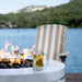 A clear glass mug filled with an amber beverage sits on a white round fire pit table outdoors, with flames visible and a striped patio chair and blue water in the background.