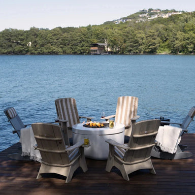 Outdoor patio furniture set on a wooden dock overlooking a lake, featuring a round fire pit table surrounded by six Adirondack-style chairs with striped and gray cushions, with a wooded shoreline in the background.