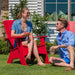 A man and a woman sitting in bright red outdoor Adirondack chairs on a green lawn, having a sunny day in front of a house with white siding and a window.