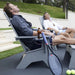 Two men relaxing in modern gray outdoor lounge chairs on a patio, with a tennis racket, bottle, and tennis balls nearby. A real outdoor environment and visible people are present, so this is Lifestyle rather than Product/Packaging.