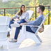 A man and a woman relax in white outdoor lounge chairs on a sunlit patio, each holding a wine glass. The scene is set against a blurred background of green foliage and a railing.