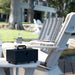 A black portable speaker with a handle sits on a white outdoor patio chair beside a drink, with striped lounge chairs and a blurred backyard setting in the background.