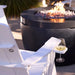 A white outdoor Adirondack chair in a patio setting with a black fire pit and flames in the background, featuring a built-in white side table on the armrest holding a wine glass.