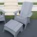 A light gray outdoor patio set featuring a high-back Adirondack chair and a matching footrest, placed on a dark gray patio with green grass and white steps in the background.