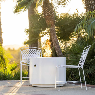 White round outdoor powdered coated fire pit and two white metal chair set on a patio garden surrounded by lush palm trees