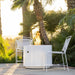 White round outdoor powdered coated fire pit and two white metal chair set on a patio garden surrounded by lush palm trees