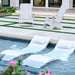 Two white outdoor lounge chairs sit on a concrete patio beside a swimming pool, with two matching white pool float loungers floating in the water in the foreground.