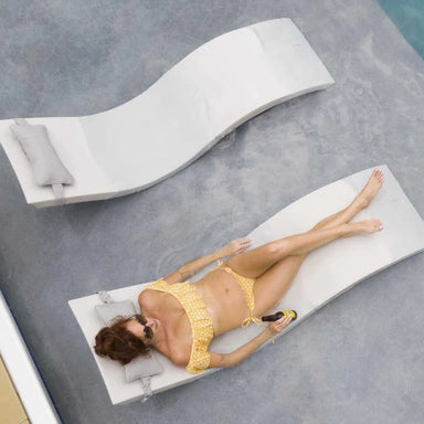 A woman in a yellow and white polka dot bikini relaxes on a white curved lounge chair next to a swimming pool, with another lounge chair visible in the background.