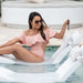 A woman wearing a light pink off-the-shoulder ruffle one-piece swimsuit and sunglasses relaxes on a white pool float in a swimming pool.