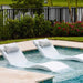 Two modern white curved outdoor chaise lounge chairs with light gray pillows, positioned in a swimming pool next to a beige stone deck, with green shrubs and a black fence in the background.