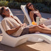 Two people reclining on modern white lounge chairs outdoors by a pool, with one person in the foreground wearing a light beige short-sleeve shirt and gray shorts and the other in a brown one-piece swimsuit. A small white side table between them holds a light green tumbler with a straw and an orange slice drink.