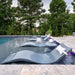 Modern curved gray outdoor lounge chairs with white pillows arranged along a swimming pool, with a stone deck and dense green trees in the background.