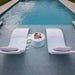Two white outdoor lounge chairs with light gray cushions are positioned on either side of a small round white side table holding a colorful magazine, all set in a swimming pool deck next to a turquoise pool.