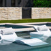 Modern white floating lounge chairs with gray pillows and a matching round side table in a swimming pool, set against a stone wall and green lawn.