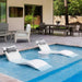 Modern outdoor poolside scene with two white curved lounge chairs, gray pillows, a small round side table, patio seating, sliding glass doors, and greenery in the background.