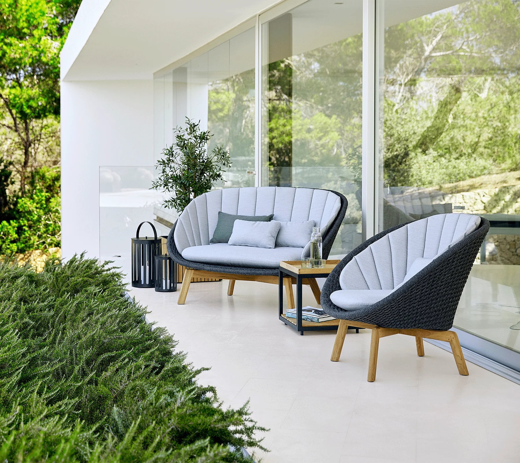  Boxhill's Peacock grey outdoor 2-seater sofa with teak base and light grey outdoor lounge chair placed beside glass wall