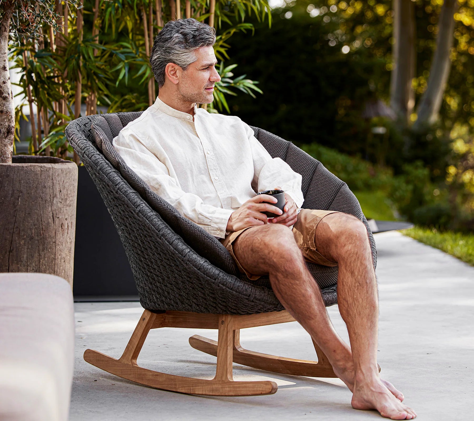 Boxhill's Peacock dark grey outdoor rocking chair with teak base with a man sitting on it holding a cup of coffee