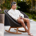 Boxhill's Peacock dark grey outdoor rocking chair with teak base with a man sitting on it holding a cup of coffee
