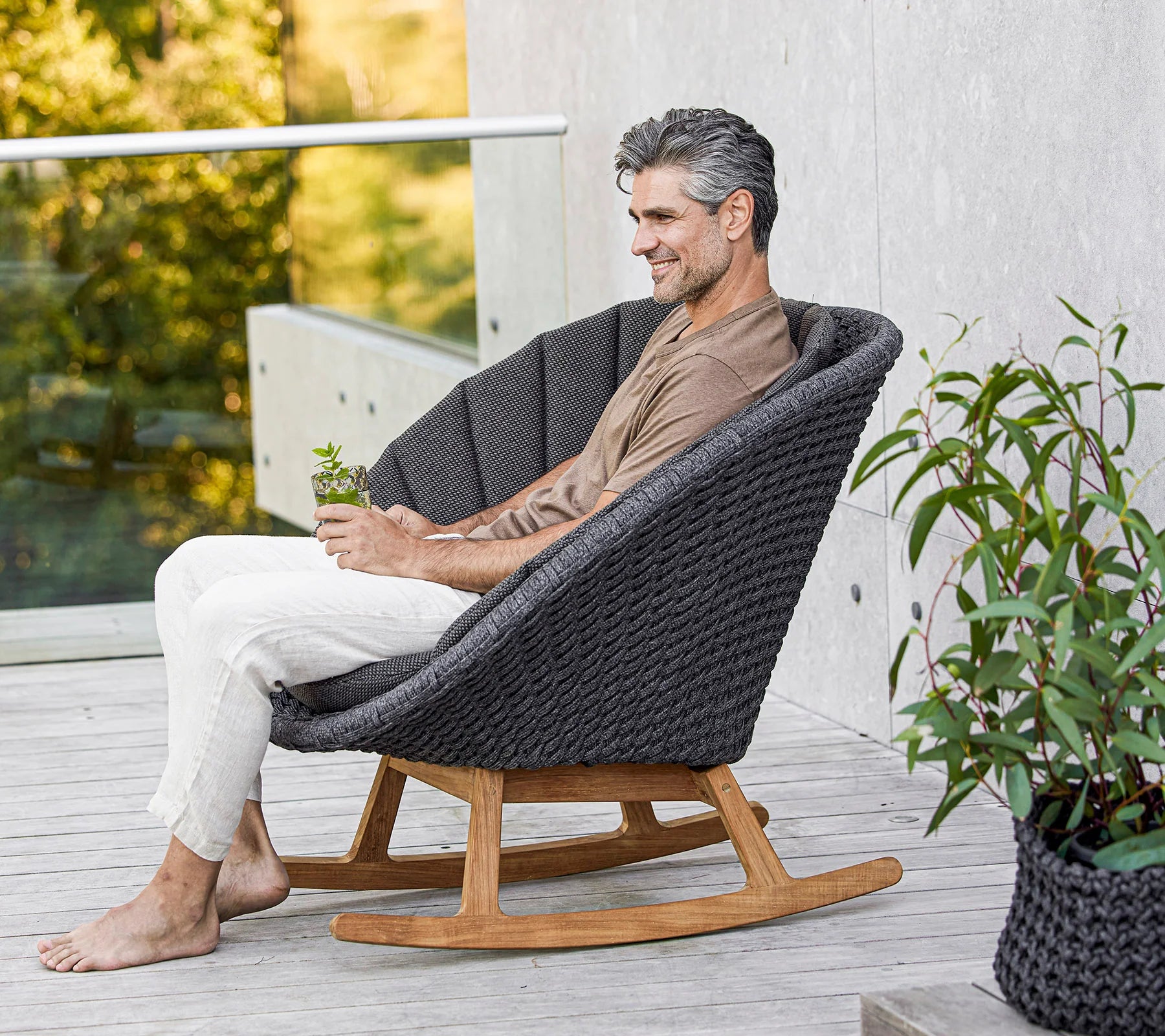  Boxhill's Peacock dark grey outdoor rocking chair with teak base with a man sitting on it holding a glass of water