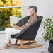  Boxhill's Peacock dark grey outdoor rocking chair with teak base with a man sitting on it holding a glass of water