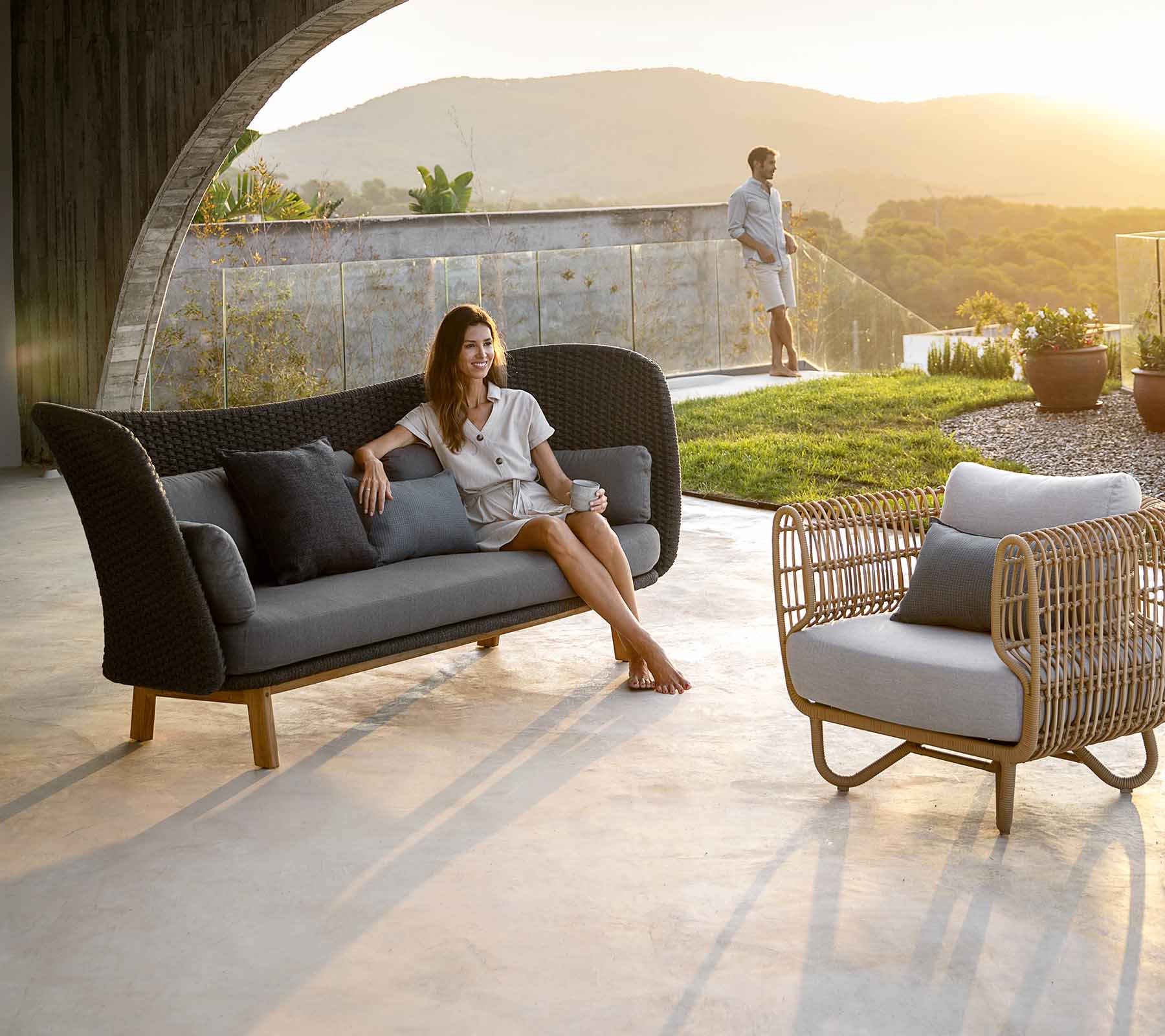 Boxhill's Peacock dark grey outdoor wing 3-seater sofa with a woman sitting on it and Nest weave outdoor lounge chair 