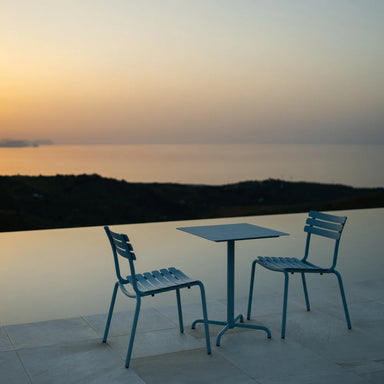 Blue square aluminum outdoor cafe table paired with two dining chairs on a tiled surface beside the pool with a view of a distant hills