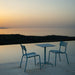 Blue square aluminum outdoor cafe table paired with two dining chairs on a tiled surface beside the pool with a view of a distant hills