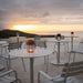 An outdoor setting with round white outdoor cafe tables and chairs illuminated by colorful table lanterns, with a sunset backdrop over hills
