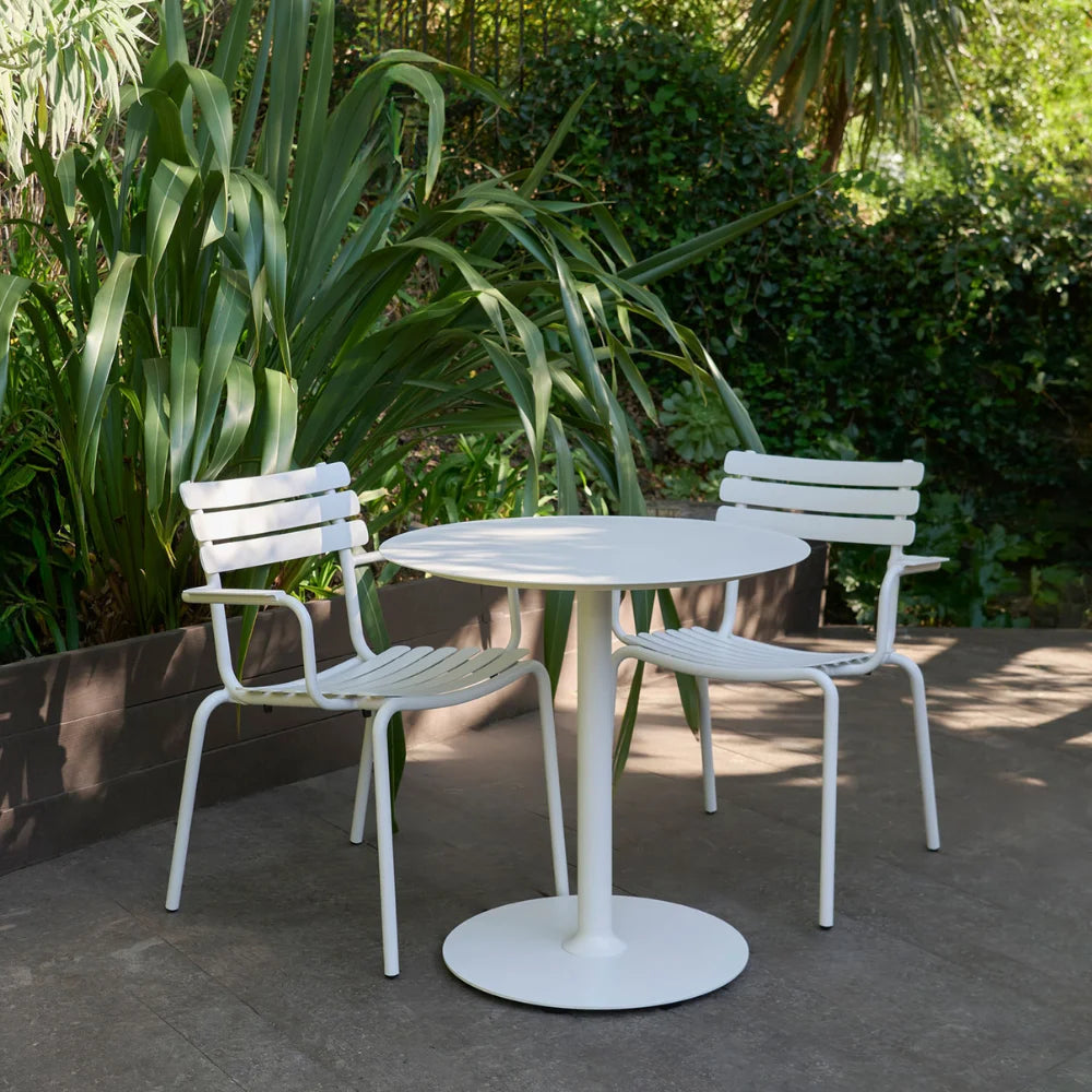 Round white aluminum outdoor cafe table paired with two metal dining chairs set on a patio surrounded with greenery