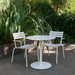 Round white aluminum outdoor cafe table paired with two metal dining chairs set on a patio surrounded with greenery