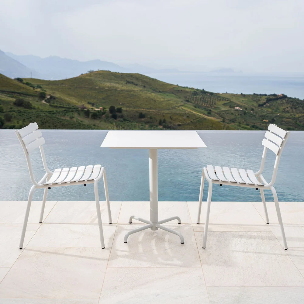 Square white aluminum patio cafe table paired with two white chairs on a tiled floor beside the pool with a view of a distant hills
