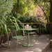 Green square metal outdoor cafe table with matching two dining chairs set on a wooden deck patio surrounded with greenery