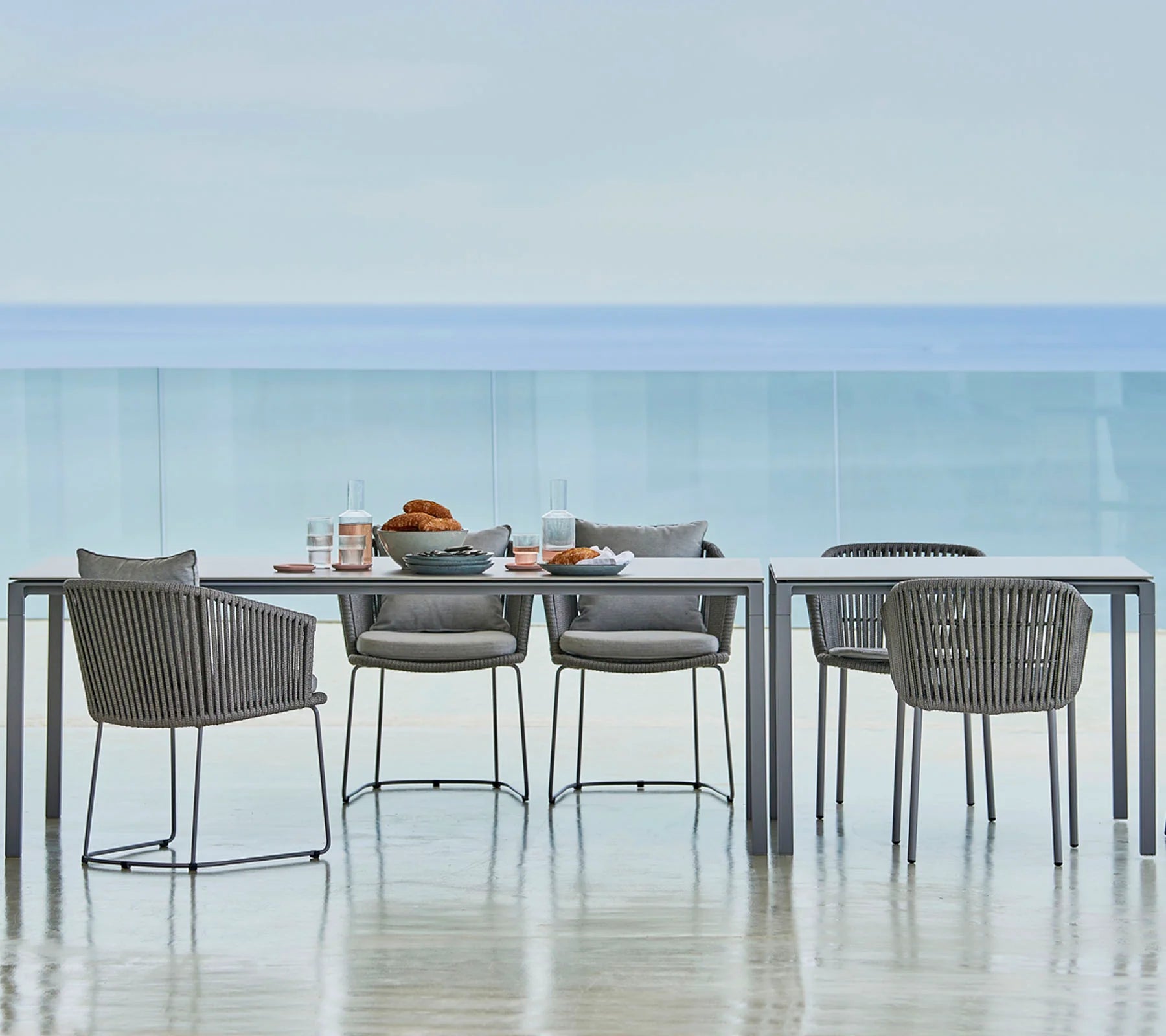 Boxhill's Pure light grey aluminum outdoor dining table with dark grey outdoor dining chairs set against ocean backdrop