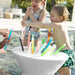 A child and an adult play with colorful plastic popsicle sticks in a white in pool side table outdoors by a swimming pool.