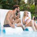 A man and a woman relax in white poolside lounge chairs by a swimming pool, with palm trees in the background. The man wears dark patterned swim trunks and holds a can of beer, while the woman wears a white bikini and sunglasses and holds an orange drink.